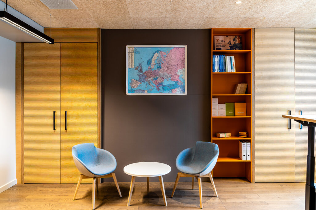 Moderni iekārtots ofiss ar skapi, plauktiem, kuros stāv grāmatas, Eiropas karti pie sienas un 2 apmeklētāju krēsliem un žurnālgaldiņu.